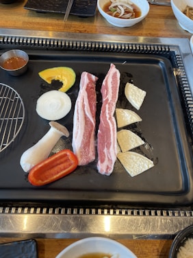 A grill with various uncooked food items including two strips of raw pork, sliced onion, squash, a king oyster mushroom, red bell pepper, and pieces of white vegetable. A small metal cup filled with sauce is on the side. There is a dish with sauce and chopped onions on the wooden table next to the grill.