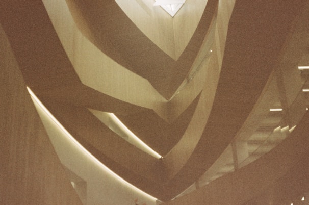 An intricate interior architectural structure featuring sweeping, curved elements with a modern design. Light filters through an upper opening, casting soft shadows and highlights on the wooden surfaces.