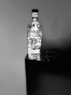 A bottle of Absolut Vodka is positioned on a dark surface, filled with small fairy lights, emitting a bright glow that highlights the text on the bottle. The background is a plain gradient from light gray to dark, casting a subtle shadow on the right side.