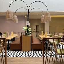 A modern indoor seating area with wooden tables and cushioned chairs. The space features a central planter with green plants and is decorated with wicker lantern-style light fixtures. The flooring has a patterned black and white design, and the area is enclosed by a rope and wood railing.