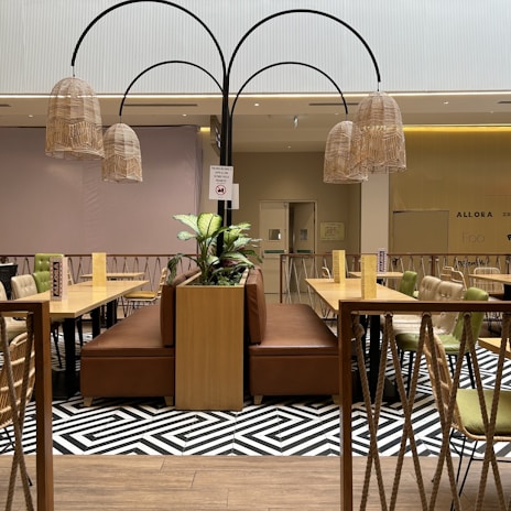 A modern indoor seating area with wooden tables and cushioned chairs. The space features a central planter with green plants and is decorated with wicker lantern-style light fixtures. The flooring has a patterned black and white design, and the area is enclosed by a rope and wood railing.