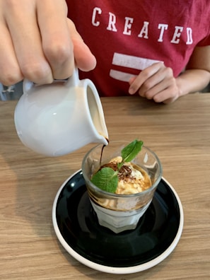 A person wearing a red shirt is pouring espresso from a small white pitcher over a glass containing a scoop of ice cream topped with fresh mint leaves. The dessert is served on a black saucer placed on a wooden table.