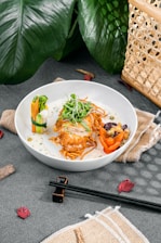 A white plate contains a finely plated dish featuring crispy fried chicken with a garnish of microgreens on top, surrounded by vegetables including broccoli, carrot, and bell pepper, as well as mixed beans. The dish is served on a bed of noodles with a creamy sauce. The setup includes chopsticks and napkins, with decorative green leaves and a wicker basket in the background.