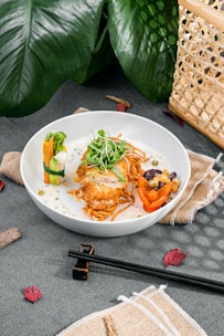 A white plate contains a finely plated dish featuring crispy fried chicken with a garnish of microgreens on top, surrounded by vegetables including broccoli, carrot, and bell pepper, as well as mixed beans. The dish is served on a bed of noodles with a creamy sauce. The setup includes chopsticks and napkins, with decorative green leaves and a wicker basket in the background.