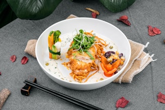 A beautifully arranged dish in a white bowl featuring a fried chicken breast on a bed of spiralized vegetables, garnished with greens. Accompaniments include colorful vegetables such as broccoli, carrot ribbons, bell pepper slices, and assorted beans. The plate is set on a gray fabric surface with decorative leaves and black chopsticks.