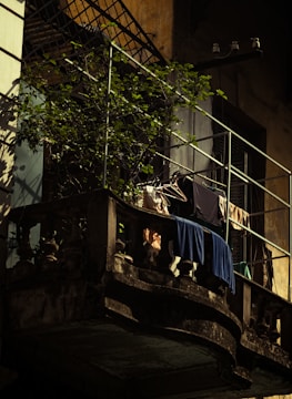 A small, rustic balcony with metal railings showcasing potted plants and clothes hanging on a line to dry. The surrounding walls have an aged appearance with some wear and discoloration. The atmosphere suggests a quiet urban setting, possibly in an older building.