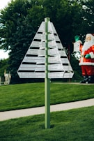 A green signpost with multiple directional signs displaying distances to various locations is set against a backdrop of lush greenery and trees. To the side, a large Santa Claus figure adds a festive touch to the scene.