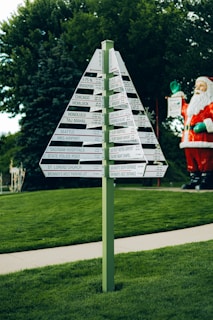 A green signpost with multiple directional signs displaying distances to various locations is set against a backdrop of lush greenery and trees. To the side, a large Santa Claus figure adds a festive touch to the scene.