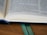 An open book with visible text on its pages, resting on a wooden surface. The book has a black cover and two green ribbons are placed as bookmarks between the pages.