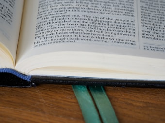 An open book with visible text on its pages, resting on a wooden surface. The book has a black cover and two green ribbons are placed as bookmarks between the pages.