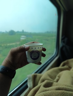 A hand holding a small paper cup with 'Indian Railways' printed on it, inside a train. The window beside shows a blurry view of greenery and open fields outside.