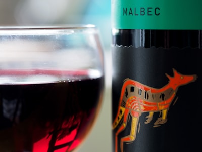 A close-up view of a glass filled with red wine and a bottle with a turquoise label featuring an illustration of an orange animal. The word 'Malbec' is printed on the label.