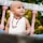 A young child with a shaved head is wearing a white sleeveless shirt and a necklace. The child is looking slightly upward with a curious and joyful expression. In the background, there are blurred green plants and part of a white and pink balustrade.