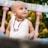 A young child with a shaved head is wearing a white sleeveless shirt and a necklace. The child is looking slightly upward with a curious and joyful expression. In the background, there are blurred green plants and part of a white and pink balustrade.