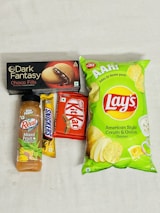 An assortment of packaged snack items including a bottle of mixed fruit juice, a Snickers bar, a Kit Kat bar, a box of Dark Fantasy Choco Fills biscuits, and a package of Lay's American Style Cream & Onion flavored chips. The items are laid out on a plain surface.