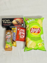An assortment of packaged snack items including a bottle of mixed fruit juice, a Snickers bar, a Kit Kat bar, a box of Dark Fantasy Choco Fills biscuits, and a package of Lay's American Style Cream & Onion flavored chips. The items are laid out on a plain surface.