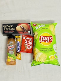 An assortment of packaged snack items including a bottle of mixed fruit juice, a Snickers bar, a Kit Kat bar, a box of Dark Fantasy Choco Fills biscuits, and a package of Lay's American Style Cream & Onion flavored chips. The items are laid out on a plain surface.
