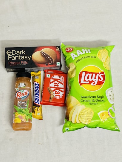 An assortment of packaged snack items including a bottle of mixed fruit juice, a Snickers bar, a Kit Kat bar, a box of Dark Fantasy Choco Fills biscuits, and a package of Lay's American Style Cream & Onion flavored chips. The items are laid out on a plain surface.