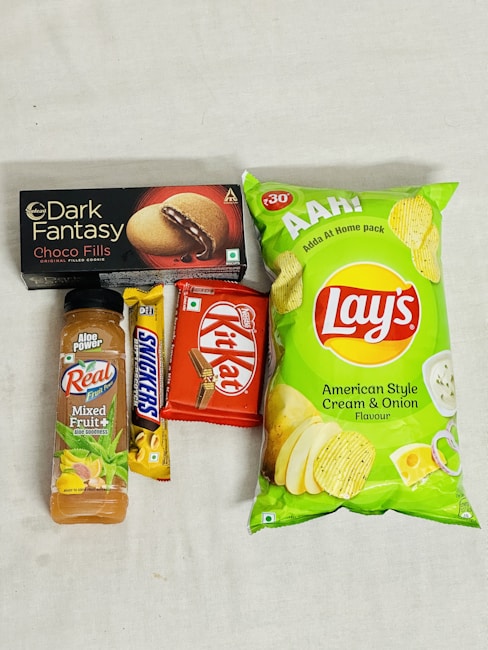 An assortment of packaged snack items including a bottle of mixed fruit juice, a Snickers bar, a Kit Kat bar, a box of Dark Fantasy Choco Fills biscuits, and a package of Lay's American Style Cream & Onion flavored chips. The items are laid out on a plain surface.