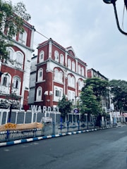 A street scene featuring a red and white colonial-style building with multiple windows and arched details. The building is adorned with signage indicating 'Ward 53' and is surrounded by trees. There is a blue and white fence along the sidewalk, and the street appears wet, suggesting recent rainfall.