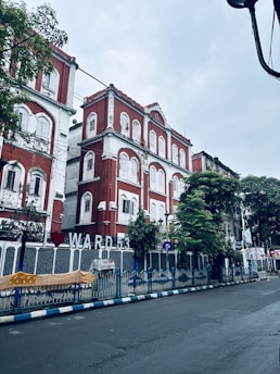 A street scene featuring a red and white colonial-style building with multiple windows and arched details. The building is adorned with signage indicating 'Ward 53' and is surrounded by trees. There is a blue and white fence along the sidewalk, and the street appears wet, suggesting recent rainfall.