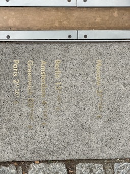 A stone pathway features engraved text indicating various city names and their corresponding longitudinal coordinates, such as Paris 2°20', Greenwich 0°00', Amsterdam 4°54', Berlin 13°25', and Moscow 37°57'. The text is inscribed in a metallic, golden color. A metal strip, secured with screws, divides the stones from a wooden platform.