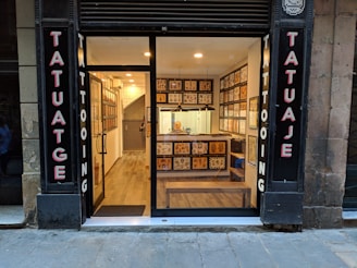 A small tattoo shop with a glass front, displaying tattoo flash art on the walls inside. The signage on either side of the entrance reads 'Tatuaje' and 'Tattooing'. Inside, a well-lit interior reveals a person seated at a counter. The shop features wooden flooring and a modern decor.