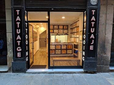 A small tattoo shop with a glass front, displaying tattoo flash art on the walls inside. The signage on either side of the entrance reads 'Tatuaje' and 'Tattooing'. Inside, a well-lit interior reveals a person seated at a counter. The shop features wooden flooring and a modern decor.