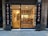 A small tattoo shop with a glass front, displaying tattoo flash art on the walls inside. The signage on either side of the entrance reads 'Tatuaje' and 'Tattooing'. Inside, a well-lit interior reveals a person seated at a counter. The shop features wooden flooring and a modern decor.