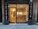 A small tattoo shop with a glass front, displaying tattoo flash art on the walls inside. The signage on either side of the entrance reads 'Tatuaje' and 'Tattooing'. Inside, a well-lit interior reveals a person seated at a counter. The shop features wooden flooring and a modern decor.