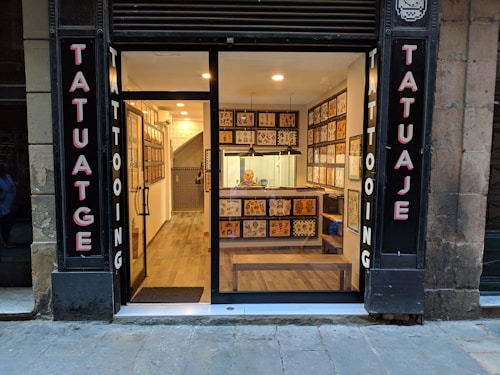 A small tattoo shop with a glass front, displaying tattoo flash art on the walls inside. The signage on either side of the entrance reads 'Tatuaje' and 'Tattooing'. Inside, a well-lit interior reveals a person seated at a counter. The shop features wooden flooring and a modern decor.