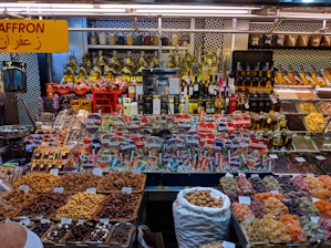 A colorful display at a market stall featuring a variety of packaged goods, spices, nuts, dried fruits, and bottles of oils and spirits. The assortment of products is neatly arranged, with saffron prominently indicated by a yellow sign in both English and Arabic. The well-lit space showcases a vibrant and organized selection, capturing the lively ambiance of a market setting.