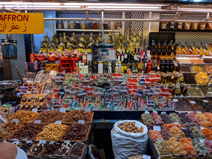 A colorful display at a market stall featuring a variety of packaged goods, spices, nuts, dried fruits, and bottles of oils and spirits. The assortment of products is neatly arranged, with saffron prominently indicated by a yellow sign in both English and Arabic. The well-lit space showcases a vibrant and organized selection, capturing the lively ambiance of a market setting.