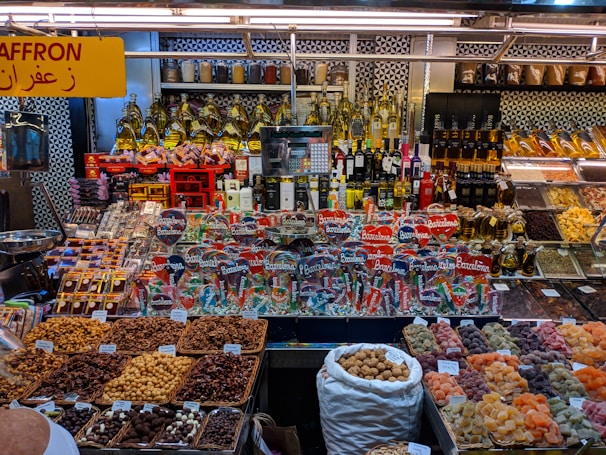 A colorful display at a market stall featuring a variety of packaged goods, spices, nuts, dried fruits, and bottles of oils and spirits. The assortment of products is neatly arranged, with saffron prominently indicated by a yellow sign in both English and Arabic. The well-lit space showcases a vibrant and organized selection, capturing the lively ambiance of a market setting.