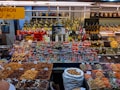 A colorful display at a market stall featuring a variety of packaged goods, spices, nuts, dried fruits, and bottles of oils and spirits. The assortment of products is neatly arranged, with saffron prominently indicated by a yellow sign in both English and Arabic. The well-lit space showcases a vibrant and organized selection, capturing the lively ambiance of a market setting.