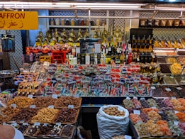 A colorful display at a market stall featuring a variety of packaged goods, spices, nuts, dried fruits, and bottles of oils and spirits. The assortment of products is neatly arranged, with saffron prominently indicated by a yellow sign in both English and Arabic. The well-lit space showcases a vibrant and organized selection, capturing the lively ambiance of a market setting.