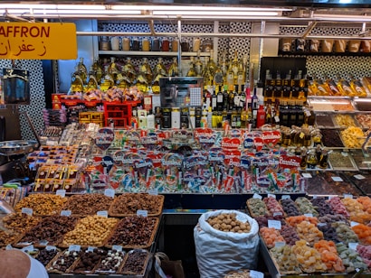 A colorful display at a market stall featuring a variety of packaged goods, spices, nuts, dried fruits, and bottles of oils and spirits. The assortment of products is neatly arranged, with saffron prominently indicated by a yellow sign in both English and Arabic. The well-lit space showcases a vibrant and organized selection, capturing the lively ambiance of a market setting.