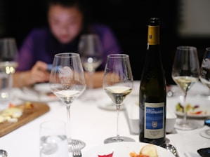 Wine glasses and a bottle of Wittmann Morstein Riesling are on a table, surrounded by plates with food remnants and a blurred person in the background using a phone. The scene suggests a post-dinner setting.