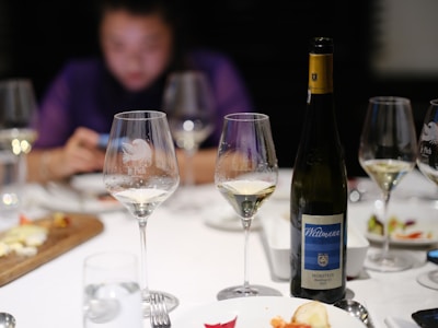 Wine glasses and a bottle of Wittmann Morstein Riesling are on a table, surrounded by plates with food remnants and a blurred person in the background using a phone. The scene suggests a post-dinner setting.