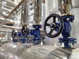 A close-up view of several industrial valves with blue metal bodies and black hand wheels on a series of pipes. The setting appears to be within a mechanical or industrial facility, with stainless steel pipes and fittings.