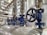 A close-up view of several industrial valves with blue metal bodies and black hand wheels on a series of pipes. The setting appears to be within a mechanical or industrial facility, with stainless steel pipes and fittings.