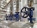 A close-up view of several industrial valves with blue metal bodies and black hand wheels on a series of pipes. The setting appears to be within a mechanical or industrial facility, with stainless steel pipes and fittings.