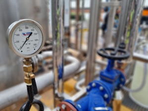 A close-up of an industrial pressure gauge displaying measurements in kilopascals with surrounding metal pipes and blue valve components in a blurred background.