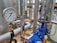 A close-up of an industrial pressure gauge displaying measurements in kilopascals with surrounding metal pipes and blue valve components in a blurred background.