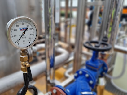 A close-up of an industrial pressure gauge displaying measurements in kilopascals with surrounding metal pipes and blue valve components in a blurred background.