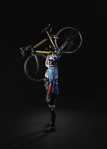 A person dressed in cycling gear holds a bicycle vertically with one hand. The individual wears a helmet, gloves, and a patterned jersey. The bike is predominantly yellow and black.