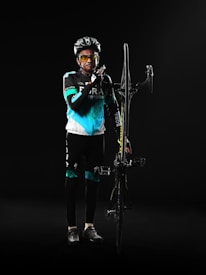 A cyclist poses with determination, wearing a black and turquoise cycling outfit. He holds a bicycle upright by the frame, with one hand resting on the handlebars. The background is dark, creating a dramatic and focused atmosphere.