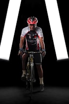 A cyclist wearing a red and black helmet, cycling gear with various logos, is positioned on a bicycle in a dimly lit environment. The background features bright white lights forming a triangular shape, enhancing the focus on the cyclist.