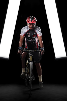 A cyclist wearing a red and black helmet, cycling gear with various logos, is positioned on a bicycle in a dimly lit environment. The background features bright white lights forming a triangular shape, enhancing the focus on the cyclist.