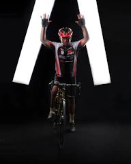 A cyclist wearing a red and white cycling jersey and helmet poses on a bicycle with both arms raised. The background is dark with bright white lights forming an inverted V shape behind the cyclist.